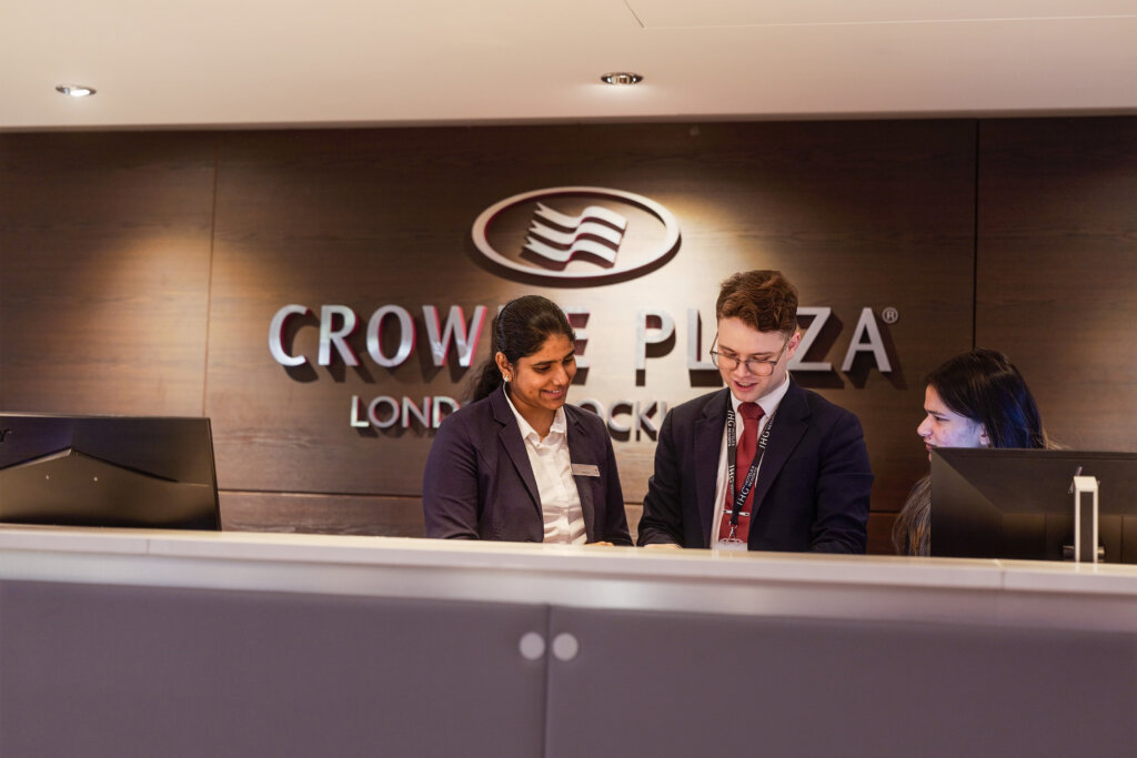 Two female employees are standing at the front desk of the Crowne Plaza London Docklands, learning something on the computer from a man in glasses.