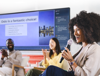 A woman with curly hair smiles while holding a microphone at a team event. There is a large presentation screen behind her with 2 colleagues sat in front of it.